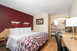 A hotel room with a bed, a desk with a chair, a mirror, and a wall-mounted light fixture.