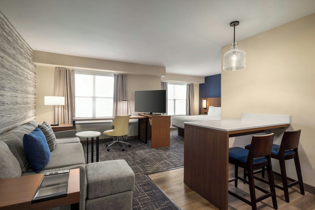 A hotel room with a sofa, a small desk with a TV, a dining area with chairs, and a large window with curtains.