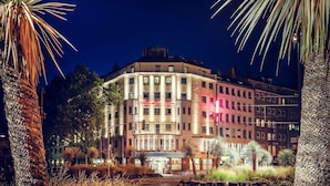 Un hotel de varios pisos con entrada central, rodeado de palmeras y una calle.