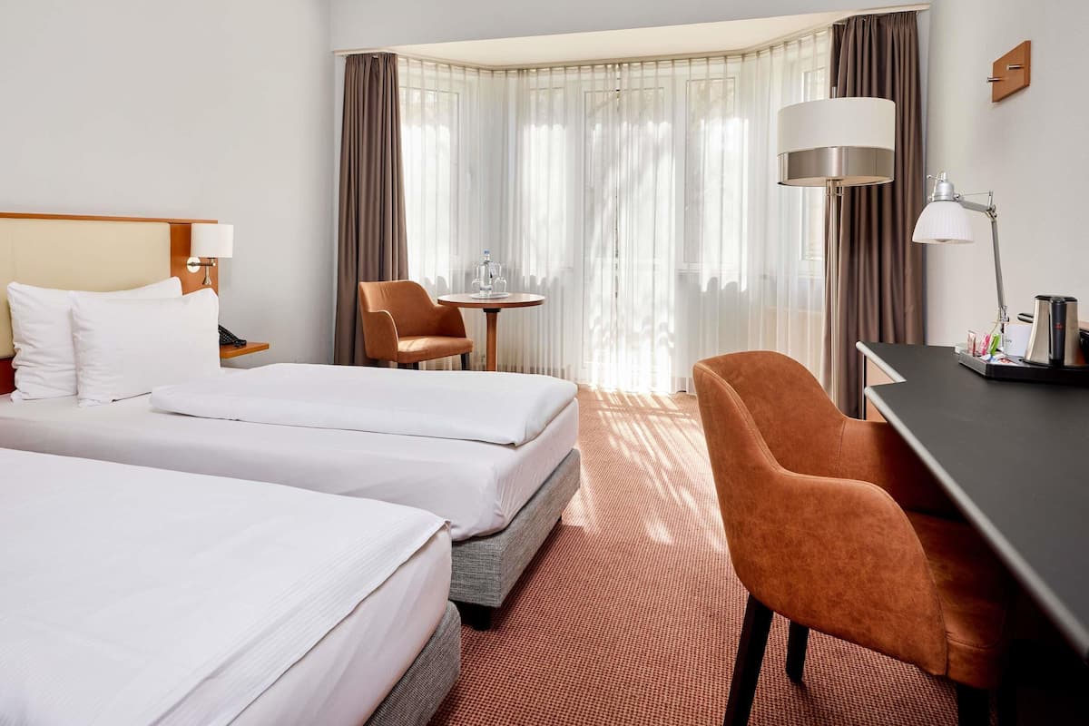A hotel room with two beds, a desk, a chair, a lamp, and a view of the outdoors.