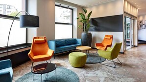 Un moderno hall de hotel con un sofá azul, sillas naranja y amarillas, una planta grande en maceta y una lámpara de pie negra.