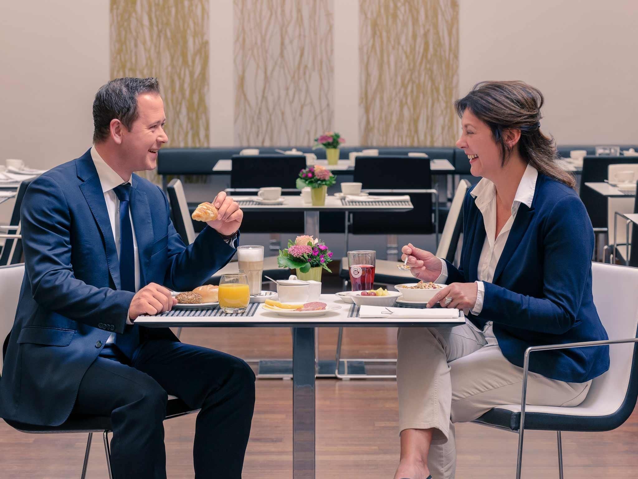 Two people seated at a table with breakfast items, a vase with flowers, and a modern interior design.