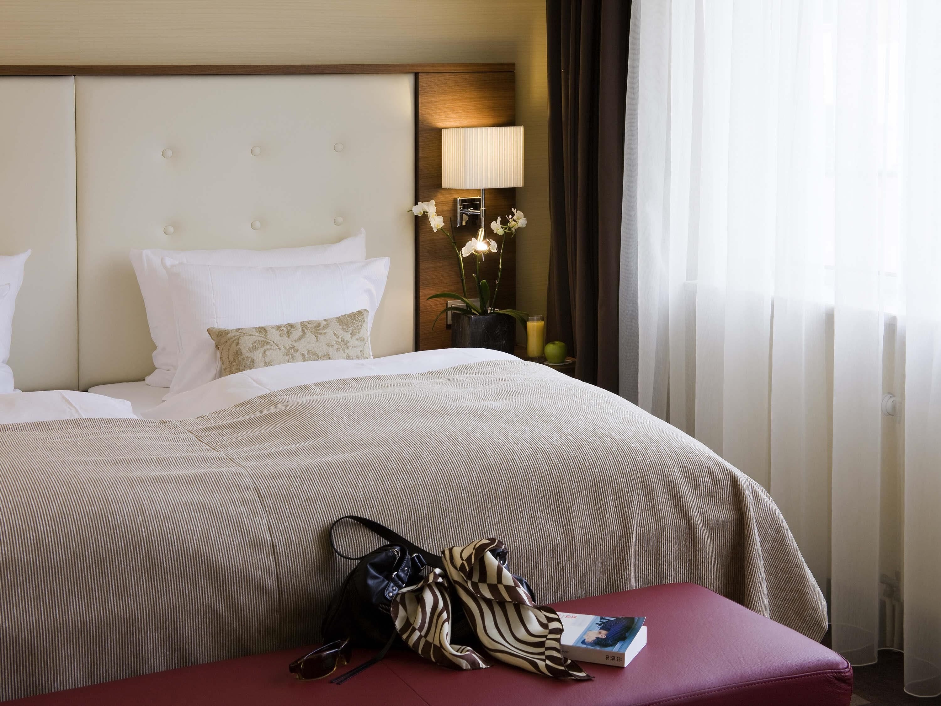 A hotel room with a bed, a bedside lamp, a vase with flowers, and a window with curtains.