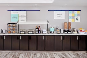 Disposição de buffet com estações de comida identificadas, máquina de café e uma cesta de frutas.