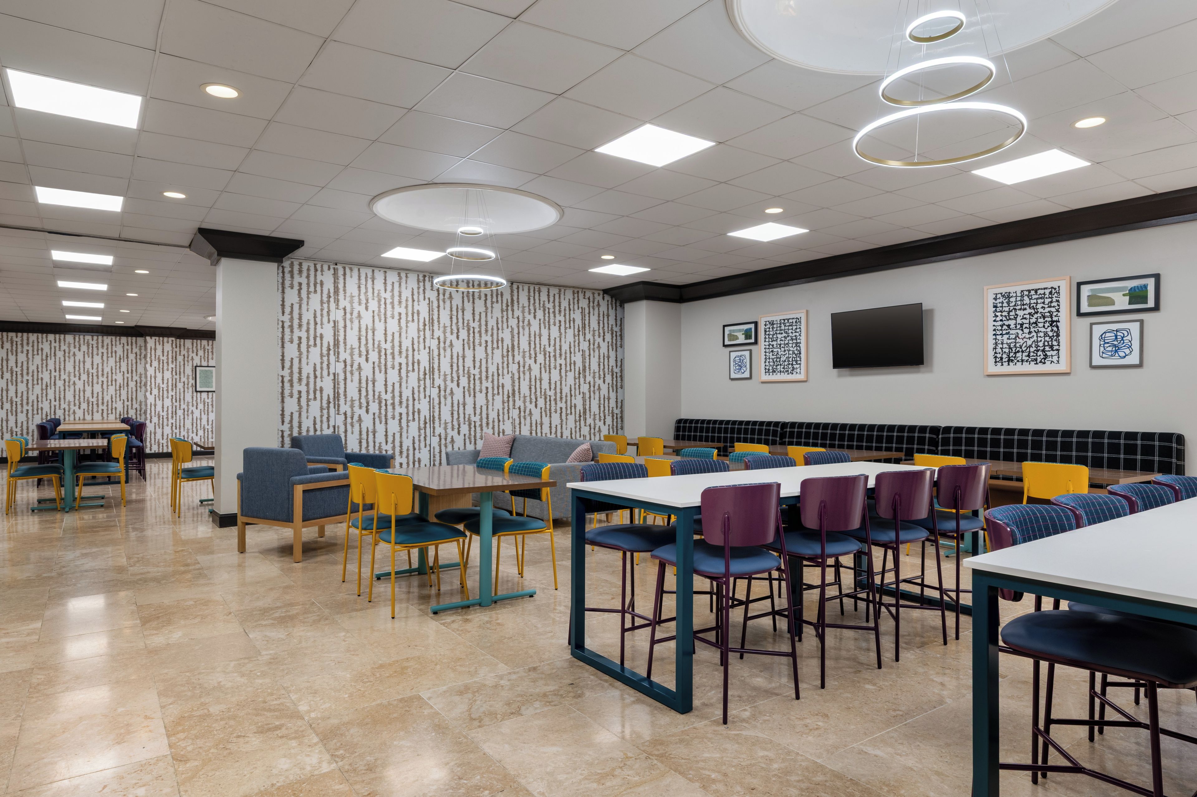 A modern dining area with multiple tables and chairs, a flat-screen TV mounted on the wall, and a patterned wallpaper design.