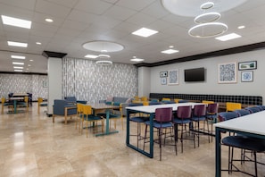 A modern dining area with multiple tables and chairs, a flat-screen TV mounted on the wall, and a patterned wallpaper design.