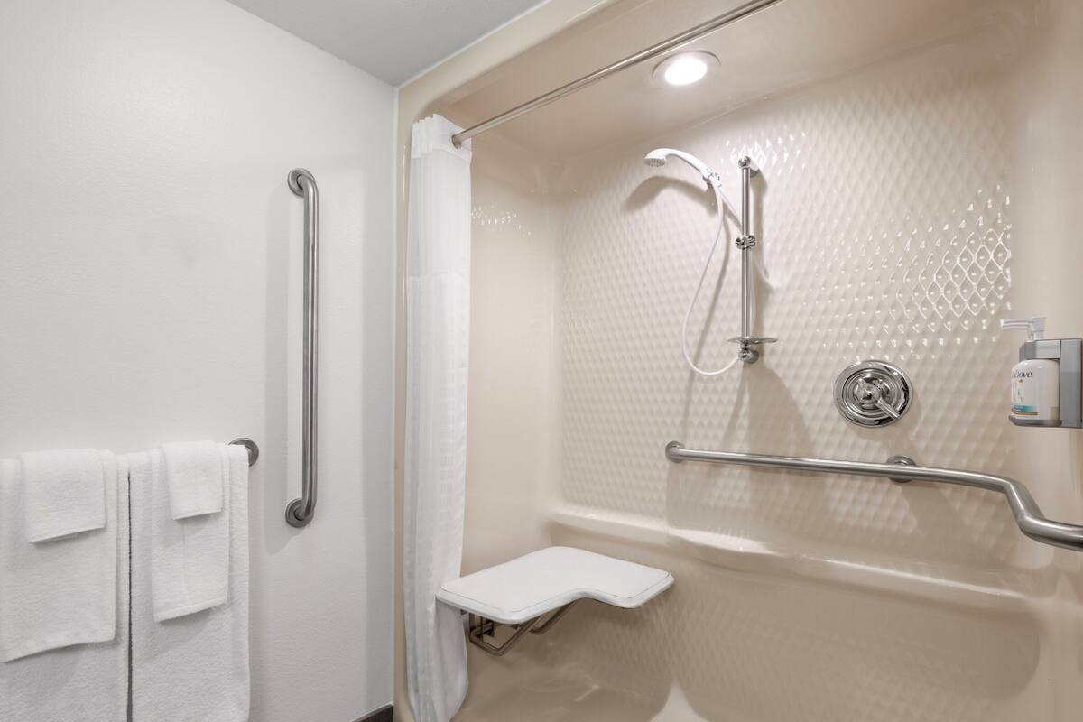 A modern bathroom with a walk-in shower, grab bars, a white bench, and a handheld showerhead.