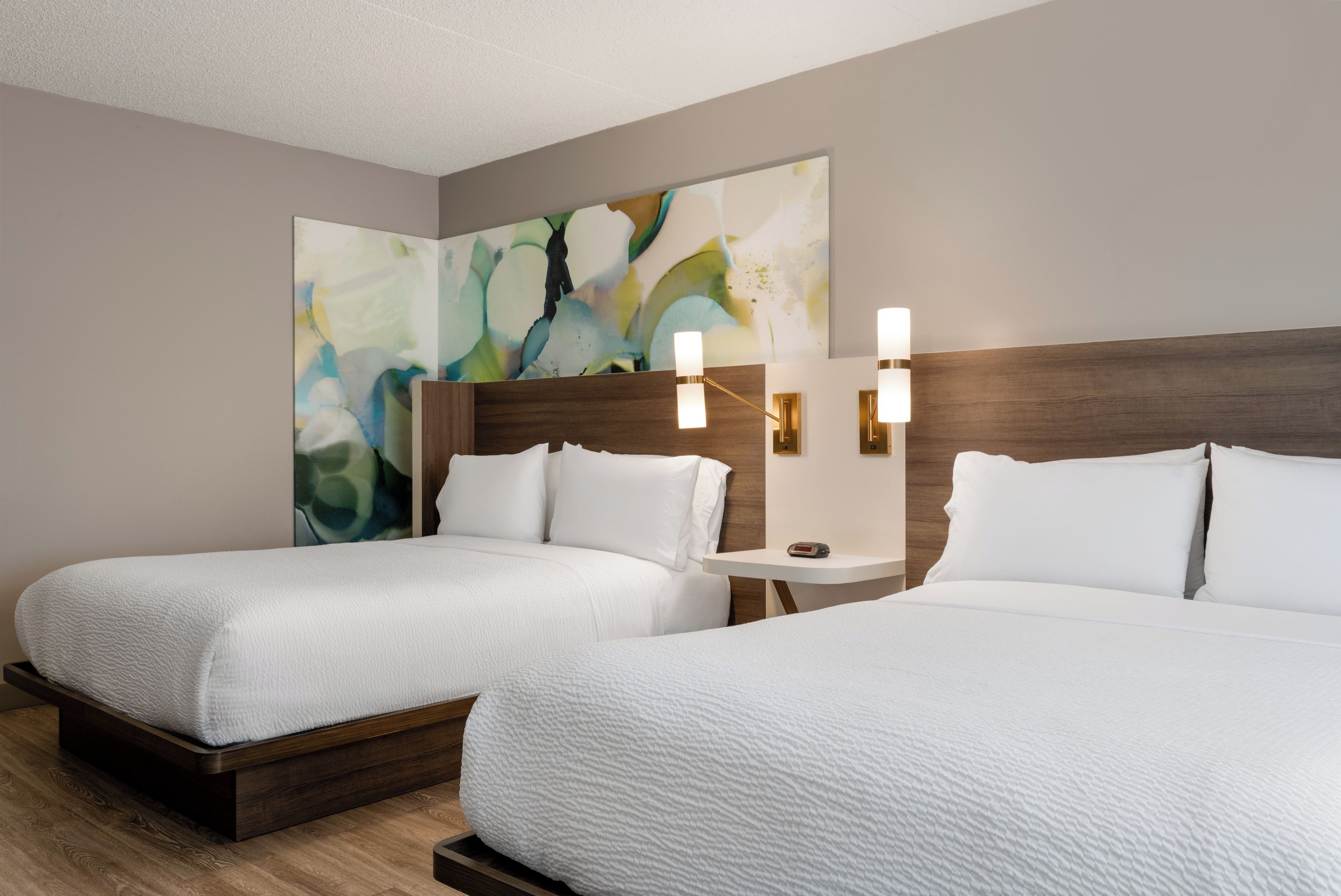 A hotel room with two beds, a large abstract painting on the wall, and a small table with two wall-mounted lights.