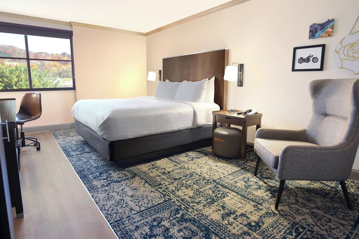 A hotel room with a large bed, a desk, a chair, and a view of autumn foliage through the window.