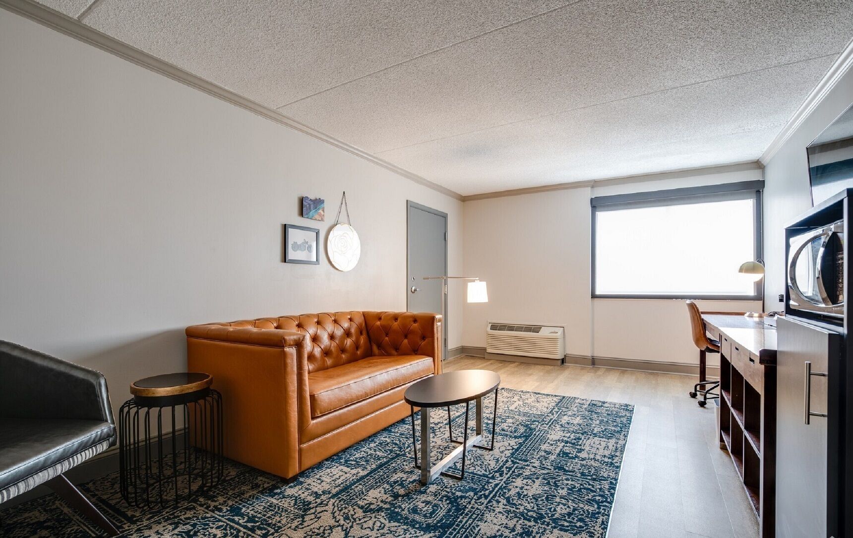 A compact living space with a brown leather sofa, a round coffee table, a patterned rug, a kitchenette with a microwave, and a large window providing natural light.
