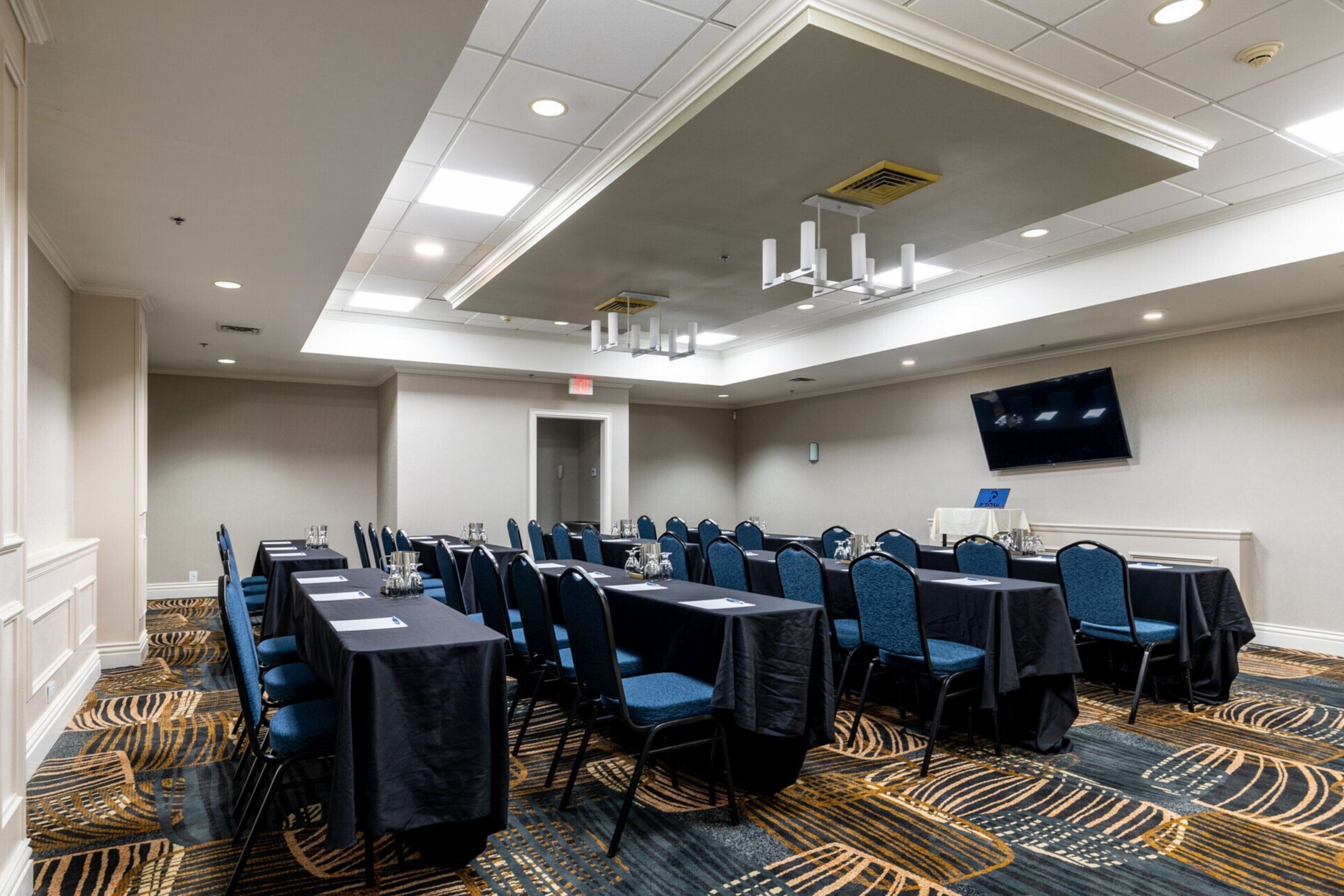 A conference room with tables and chairs, a flat-screen TV on the wall, and a patterned carpet.