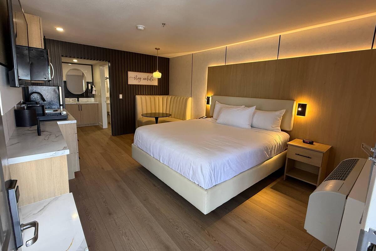 A modern hotel room with a large bed, a bedside table, a desk with a chair, a mirror, and a kitchenette.