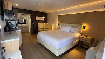 Quarto de hotel moderno com uma cama grande, uma mesa de cabeceira, escrivaninha com cadeira, espelho e cozinha compacta.