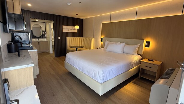 Une chambre d’hôtel moderne dotée d’un grand lit, de tables de chevet, d’un bureau avec une chaise, d’un miroir et d’un coin cuisine.