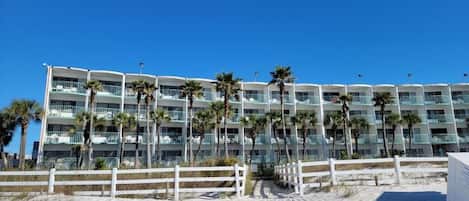 Un hotel frente a la playa con varios balcones, palmeras y una valla blanca.