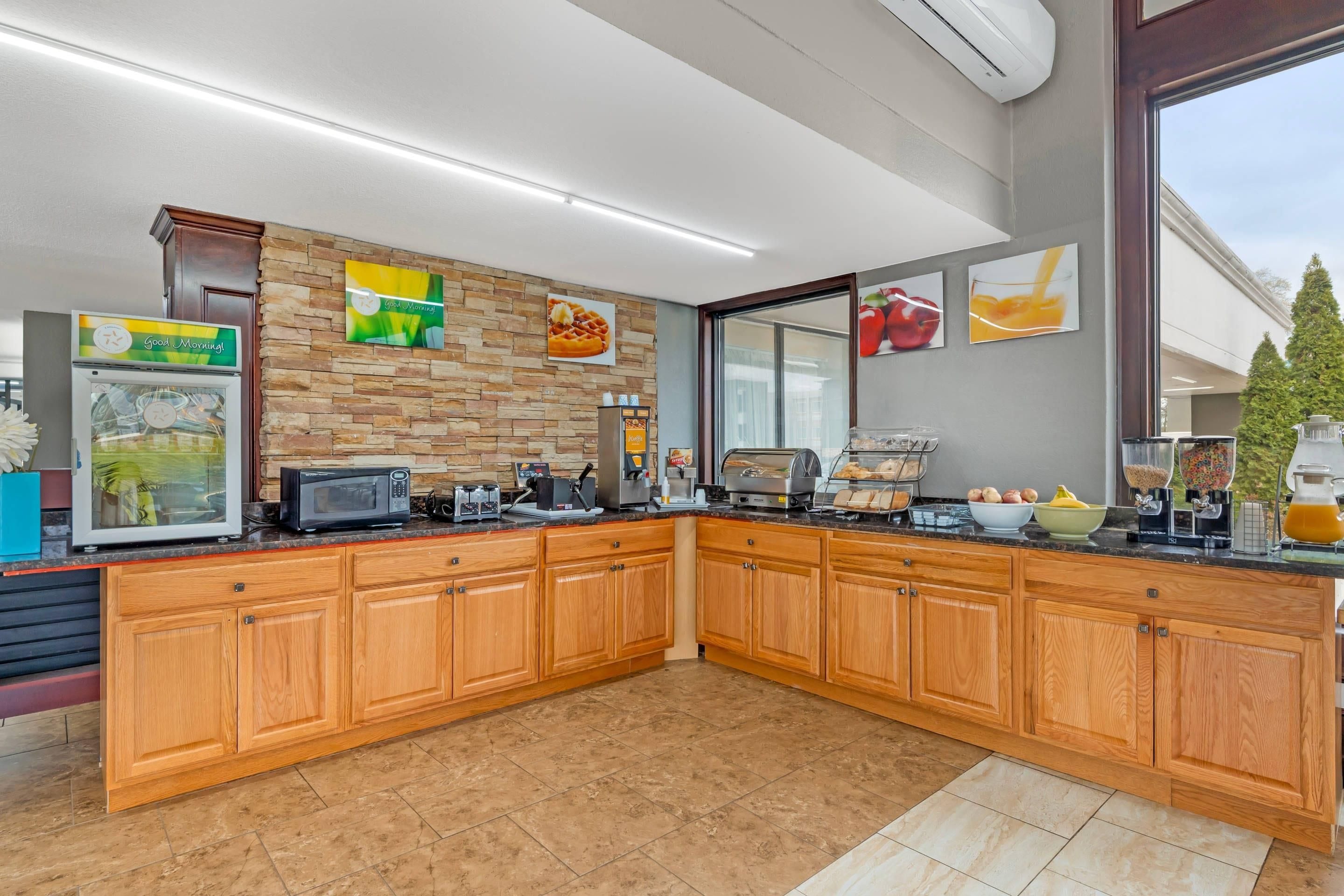 A kitchen with a stone backsplash, wooden cabinets, a microwave, a coffee machine, and a fruit display.
