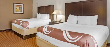 Quarto de hotel com duas camas, uma mesinha de cabeceira com abajur e um quadro na parede.