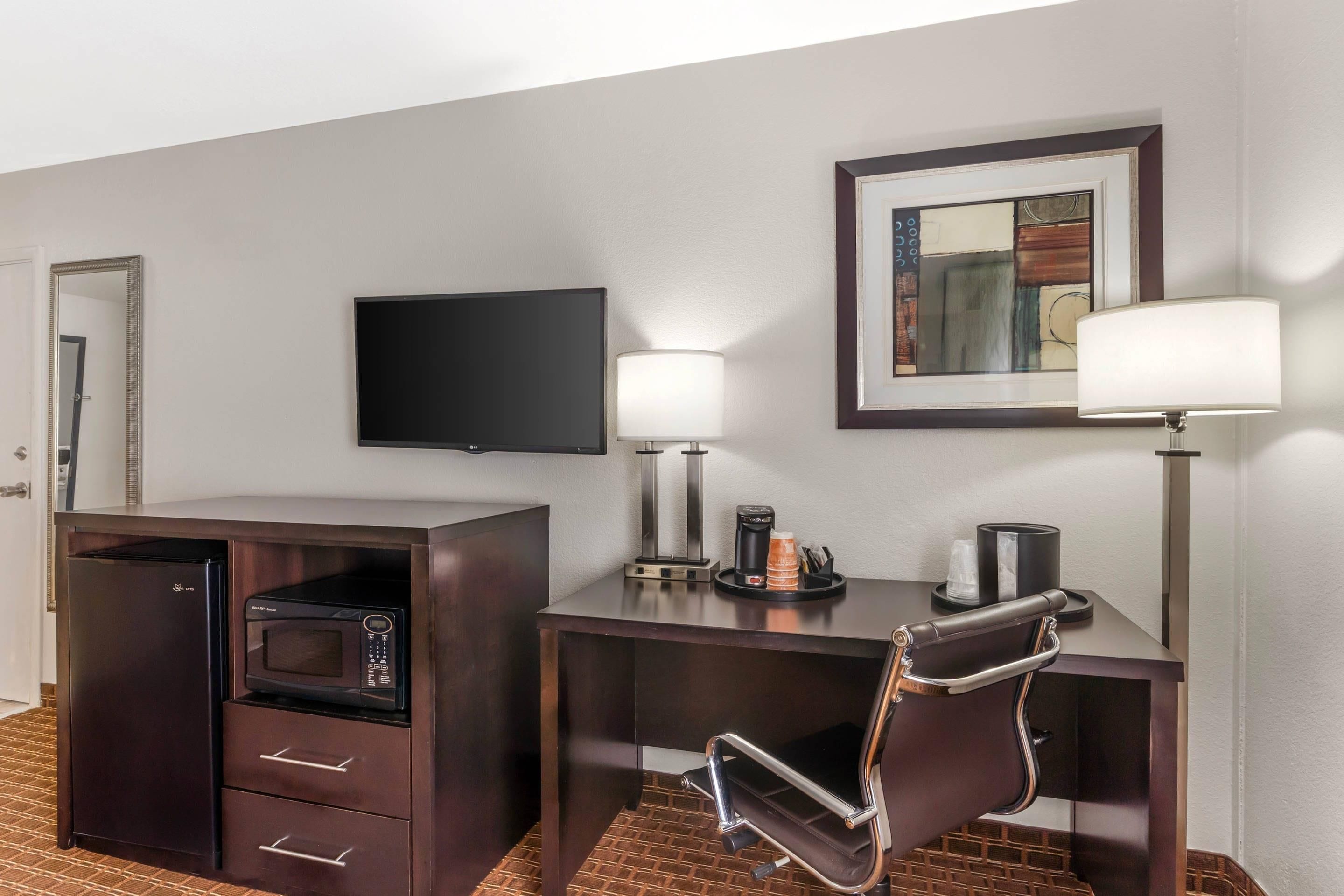 A hotel room with a dark wooden desk, a microwave, a flat-screen TV, a lamp, and a framed artwork on the wall.