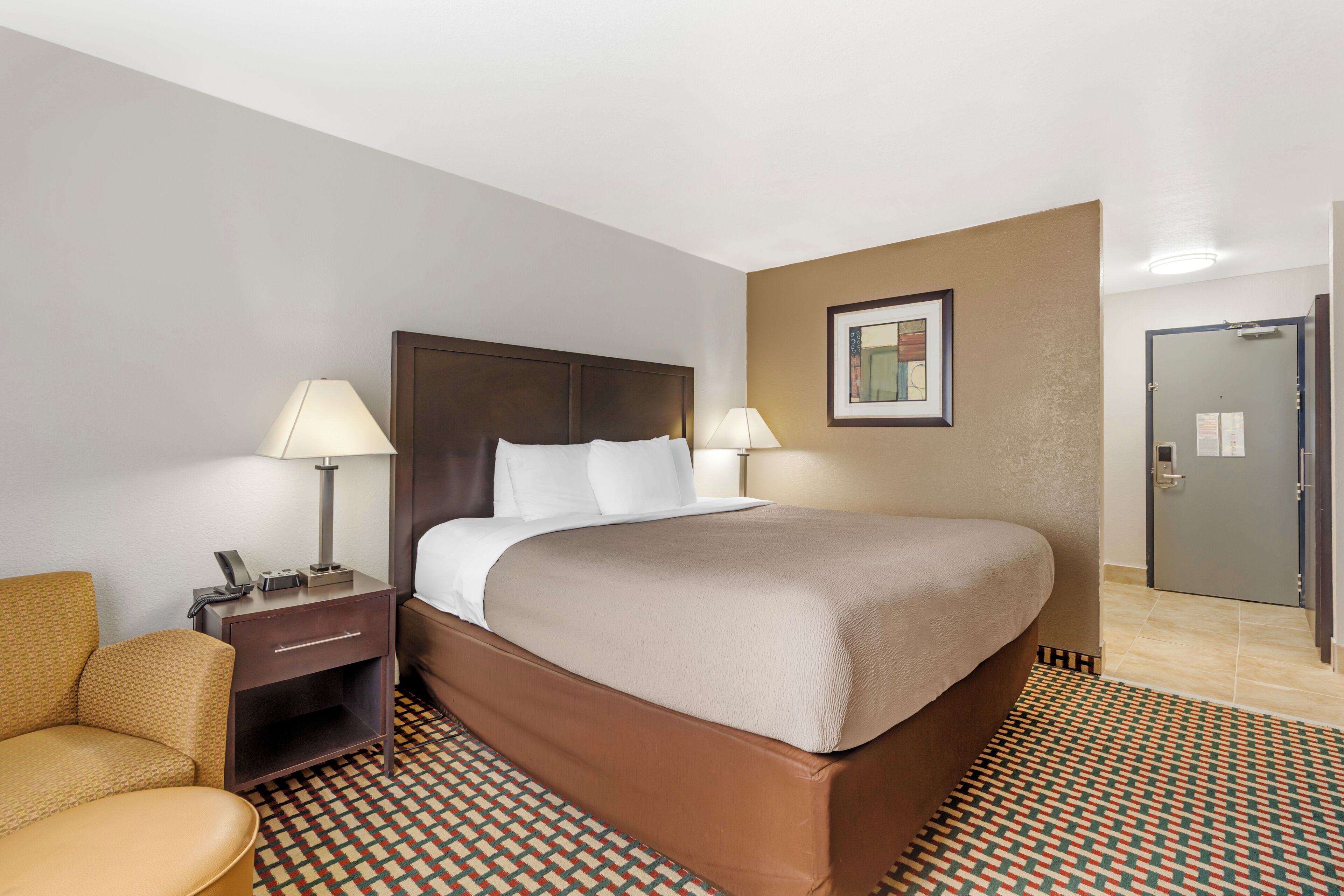 Quarto de hotel com cama, mesas de cabeceira, uma cadeira, tapete estampado e uma porta.