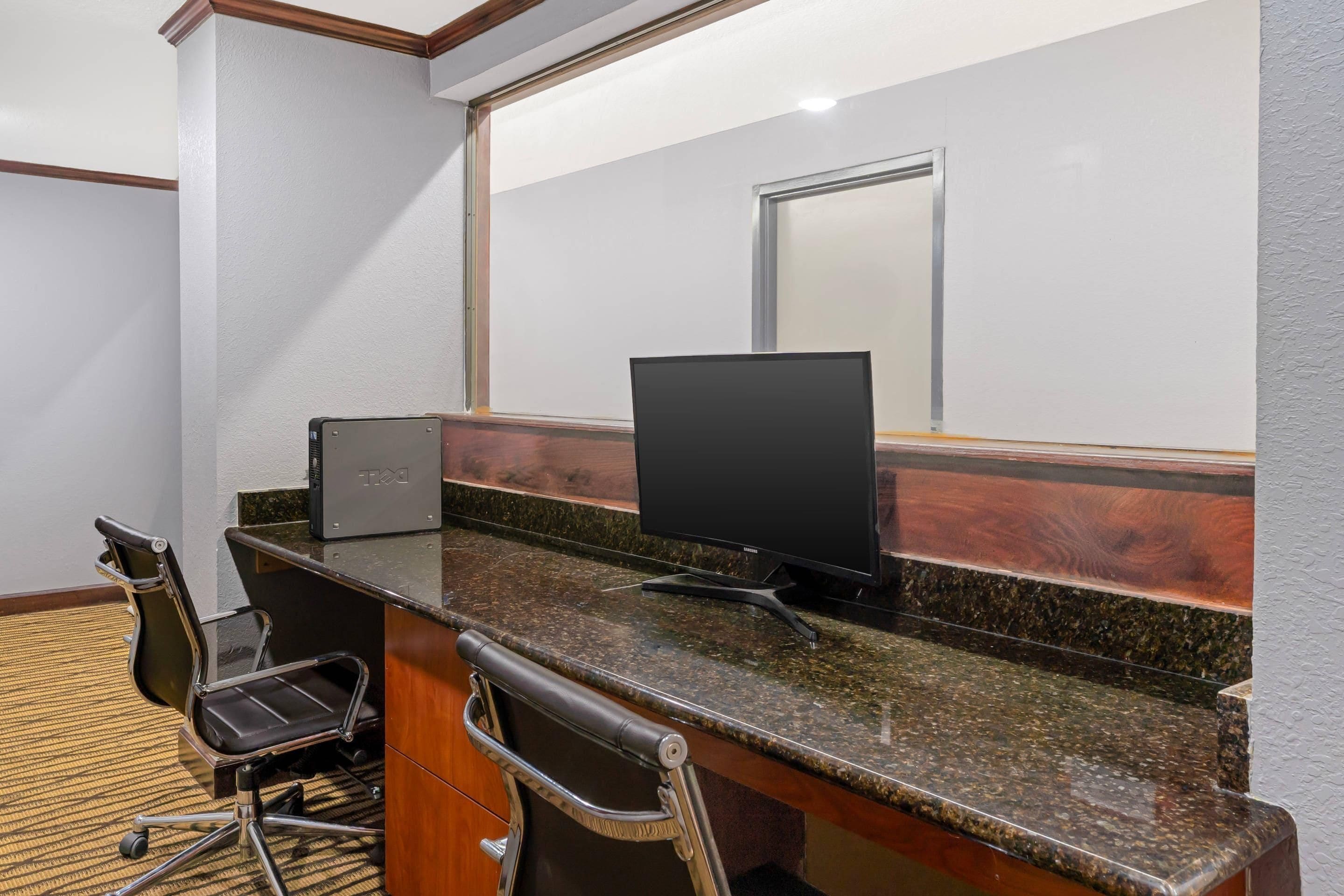 Quarto de hotel com uma mesa, duas cadeiras, um computador e uma janela grande.