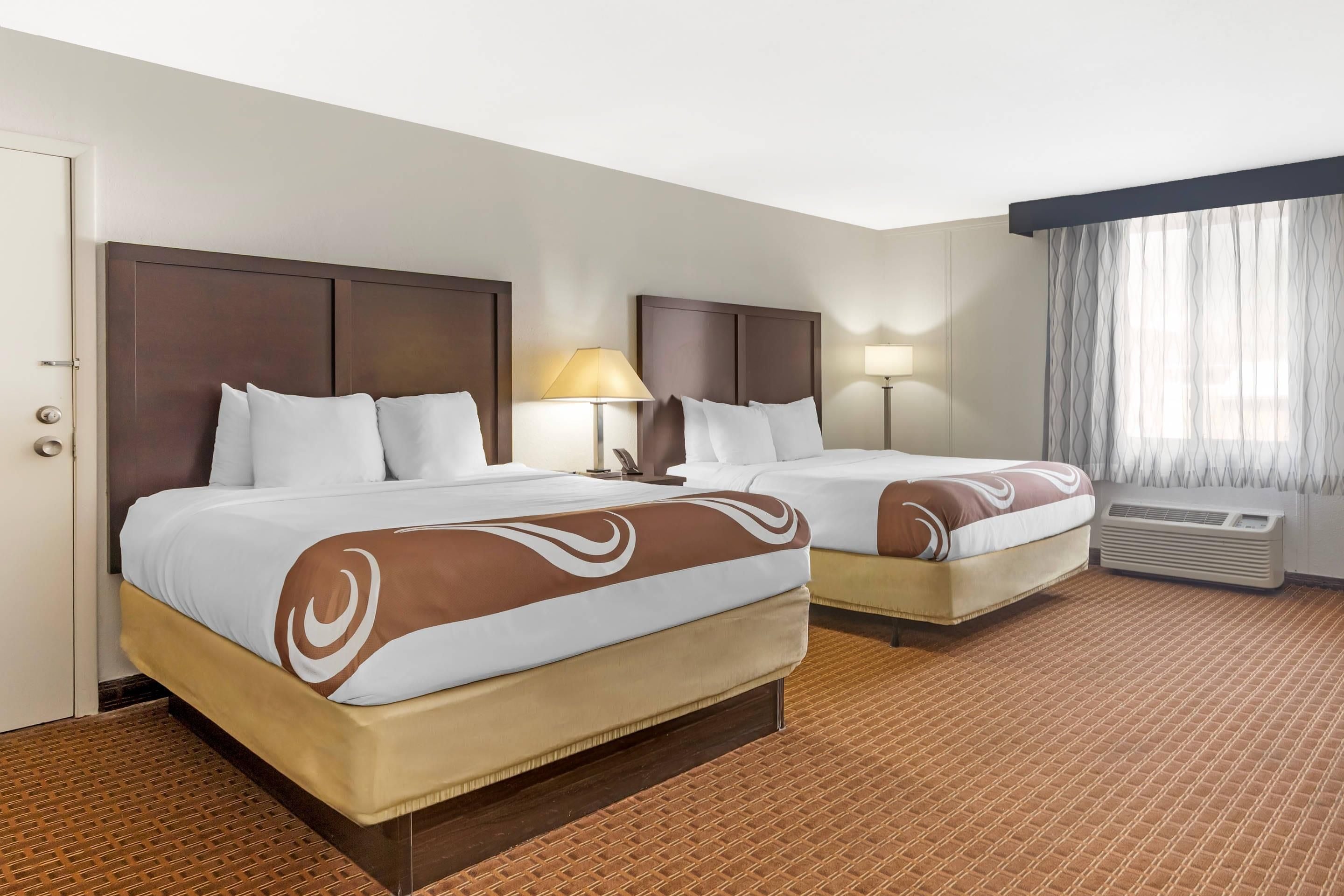 A hotel room with two beds, a door, a window with curtains, and an air conditioning unit.