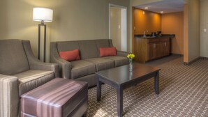 Een hotelkamer met een bank, fauteuil, poef, salontafel, lamp en kitchenette.