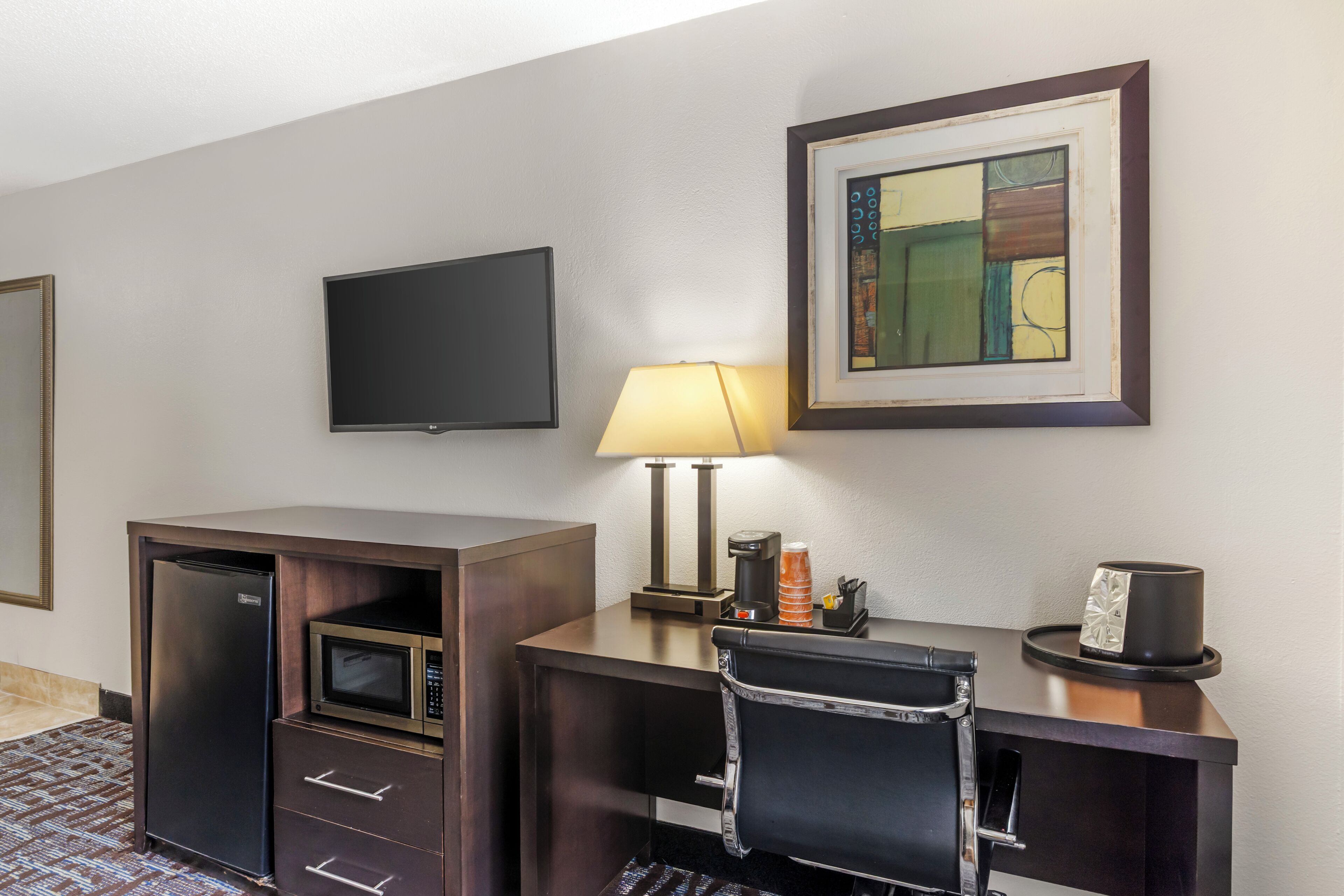 A hotel room with a desk, microwave, refrigerator, TV, lamp, and a framed artwork on the wall.