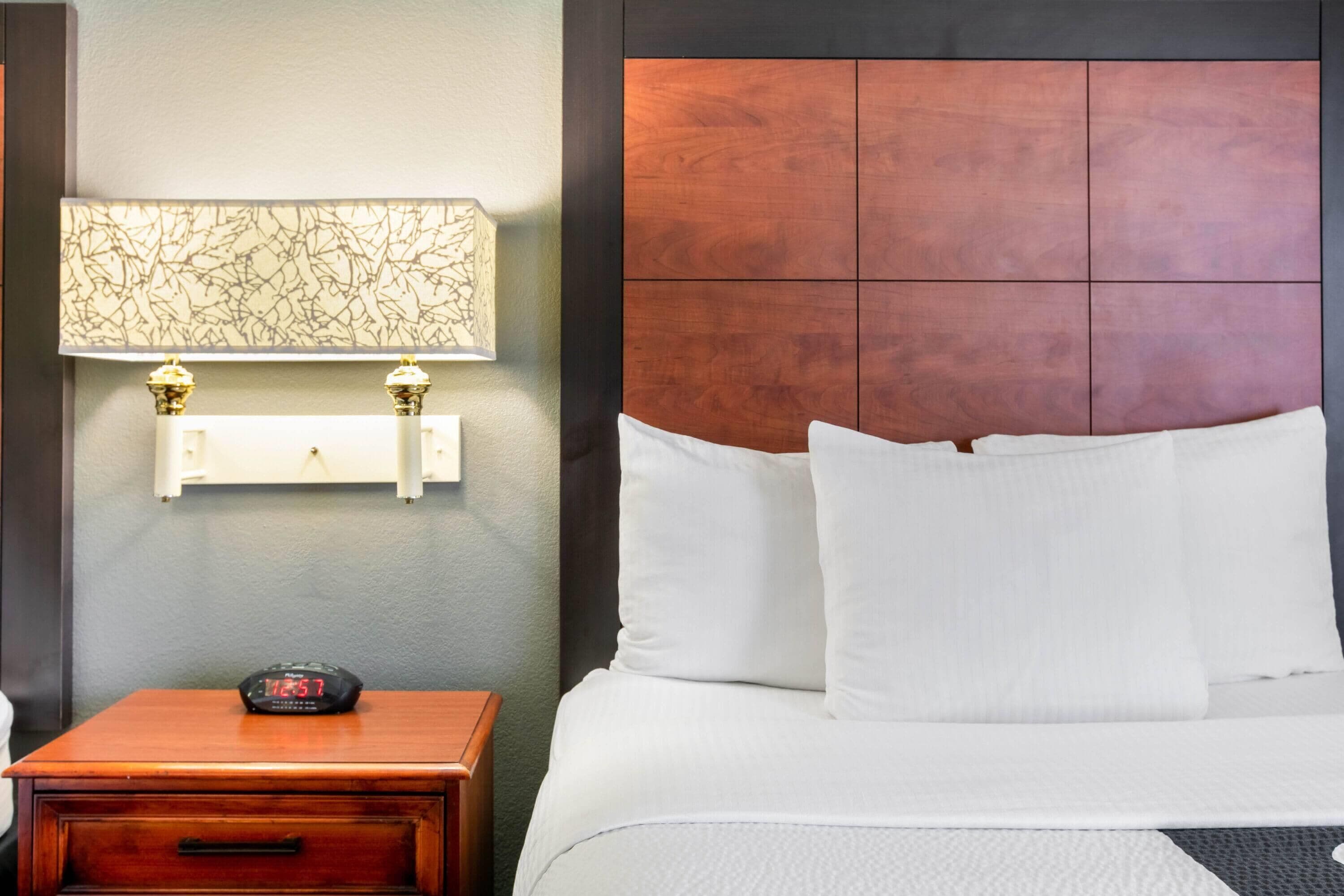 A hotel room with a wooden nightstand, a digital clock, a wall-mounted lamp, and a bed with white bedding.