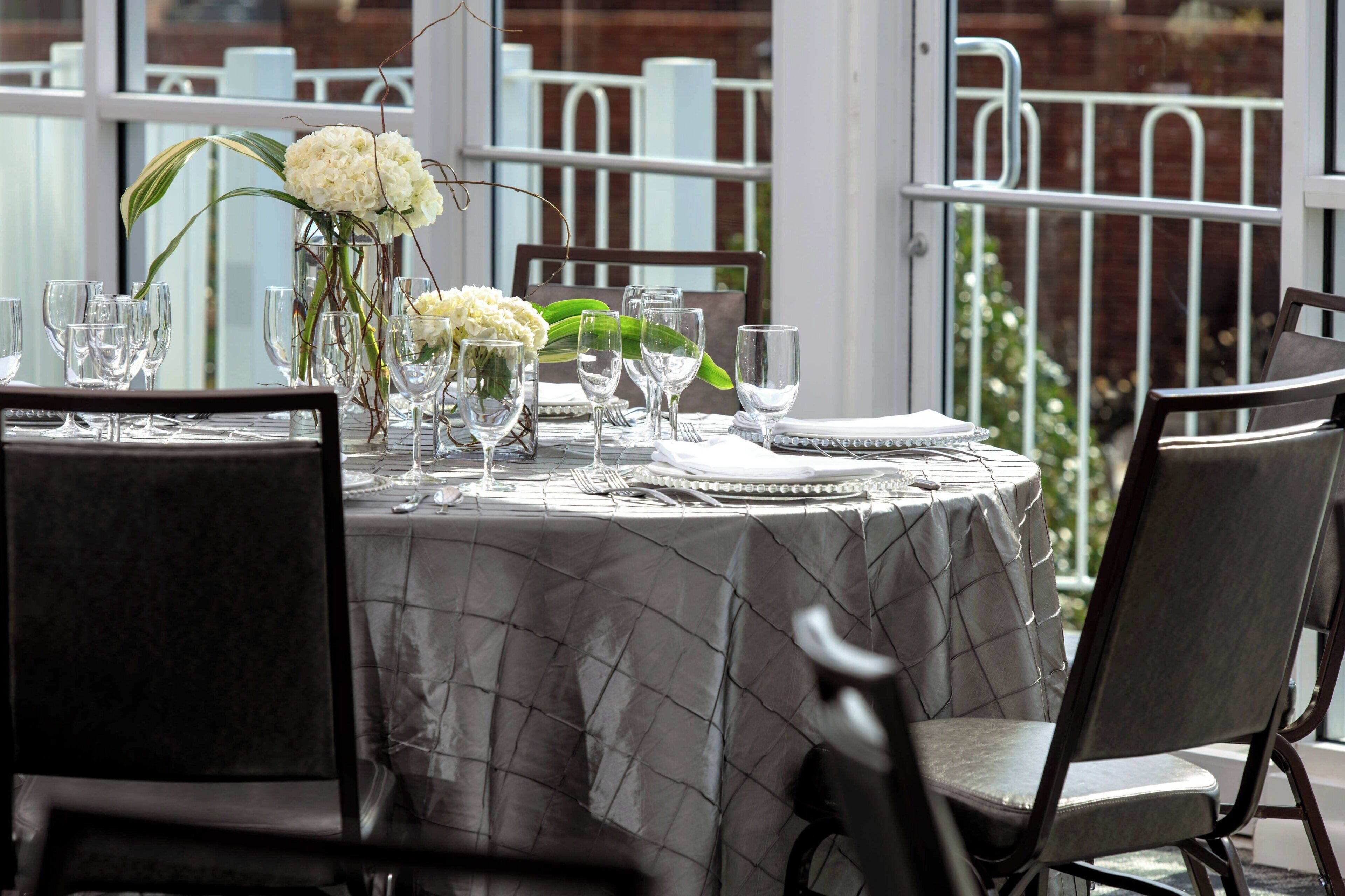 A dining table set for four with a white tablecloth, clear glassware, and a vase with white flowers.