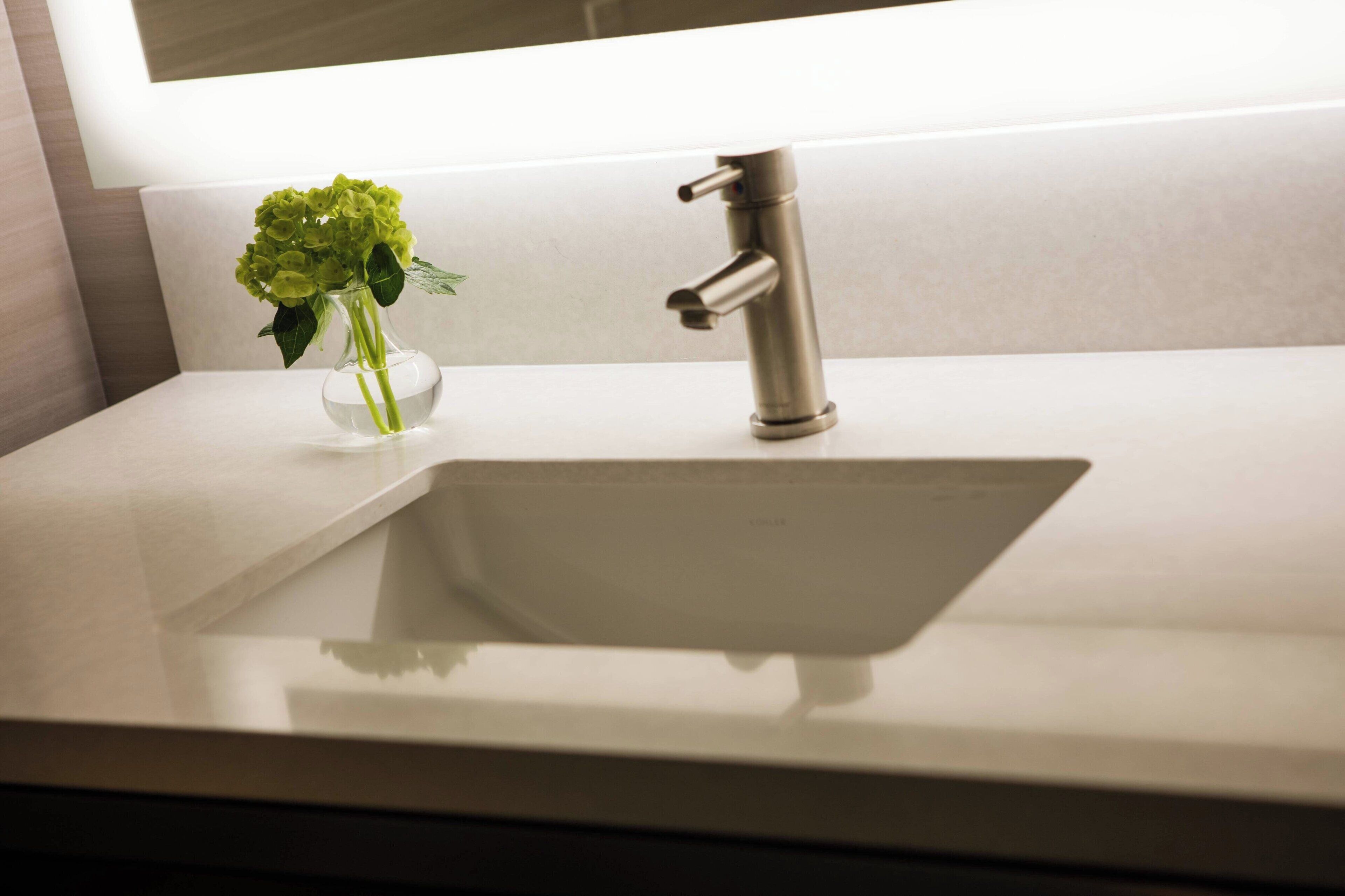 Un baño moderno con un lavamanos blanco, un solo grifo y un florero con flores verdes sobre la encimera.