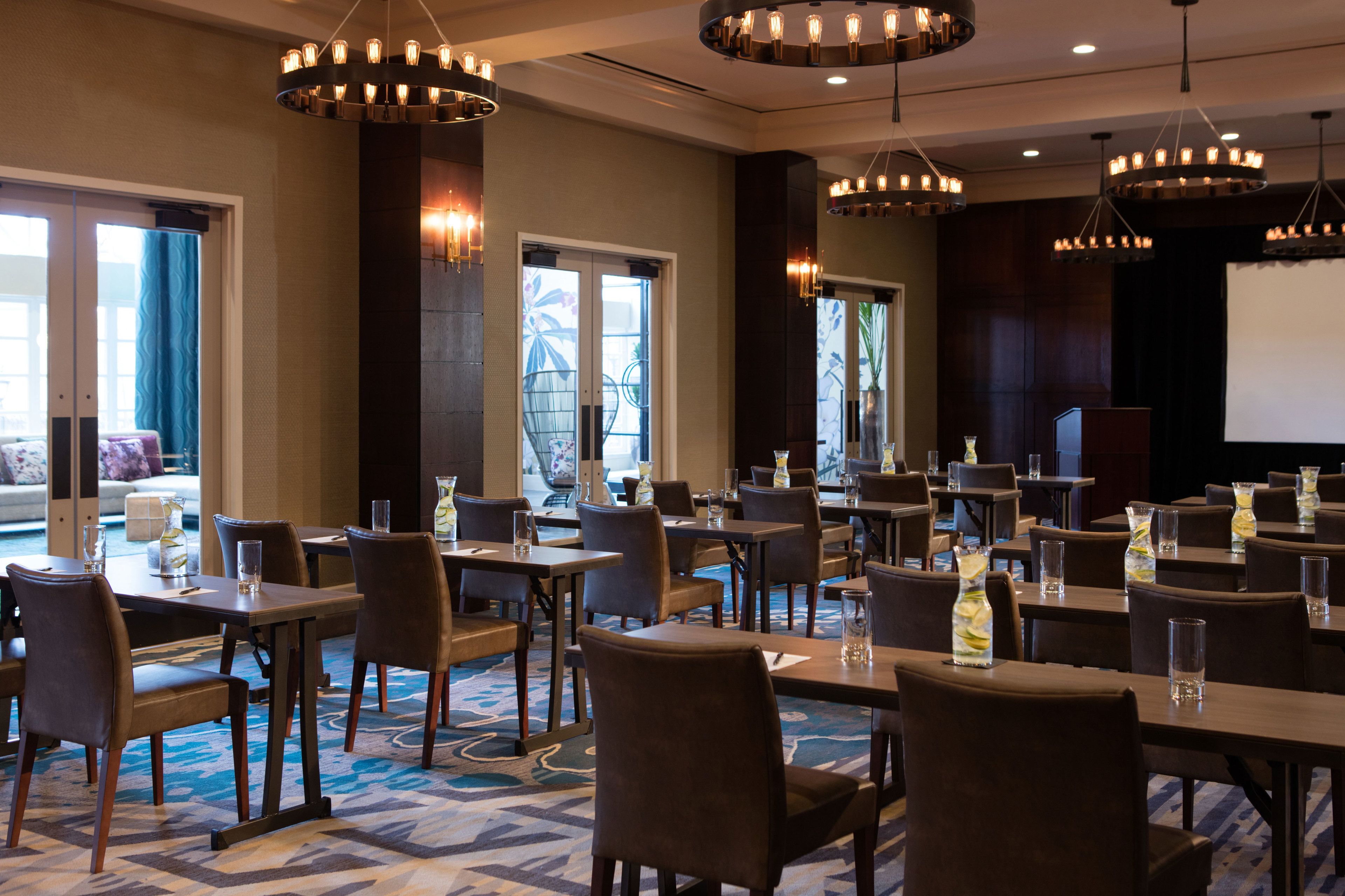 A hotel dining area with multiple tables and chairs, large windows, and chandeliers.
