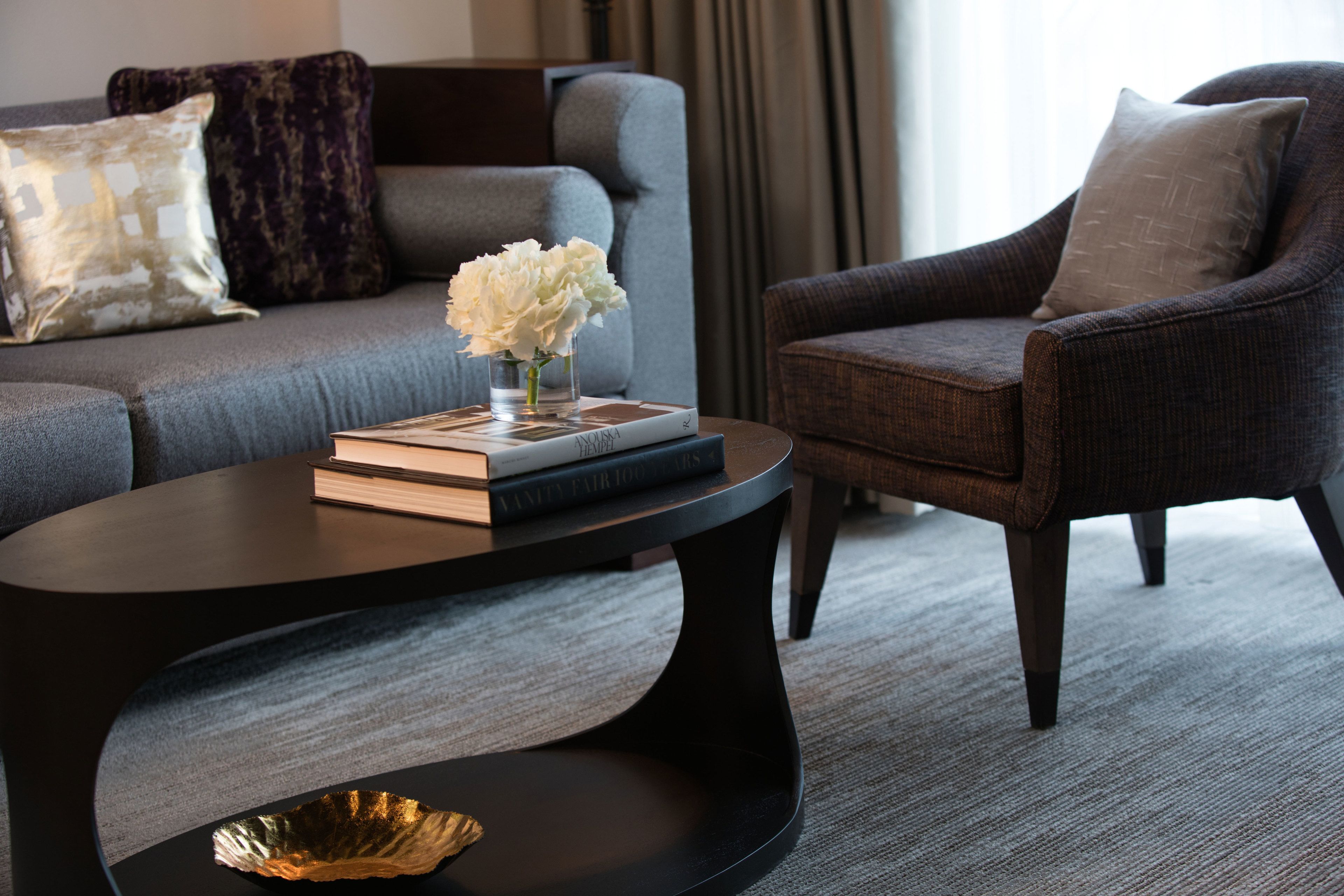 Un salon moderne avec un canapé gris, un fauteuil foncé, une table basse ronde sur laquelle se trouvent un livre et un vase de fleurs.