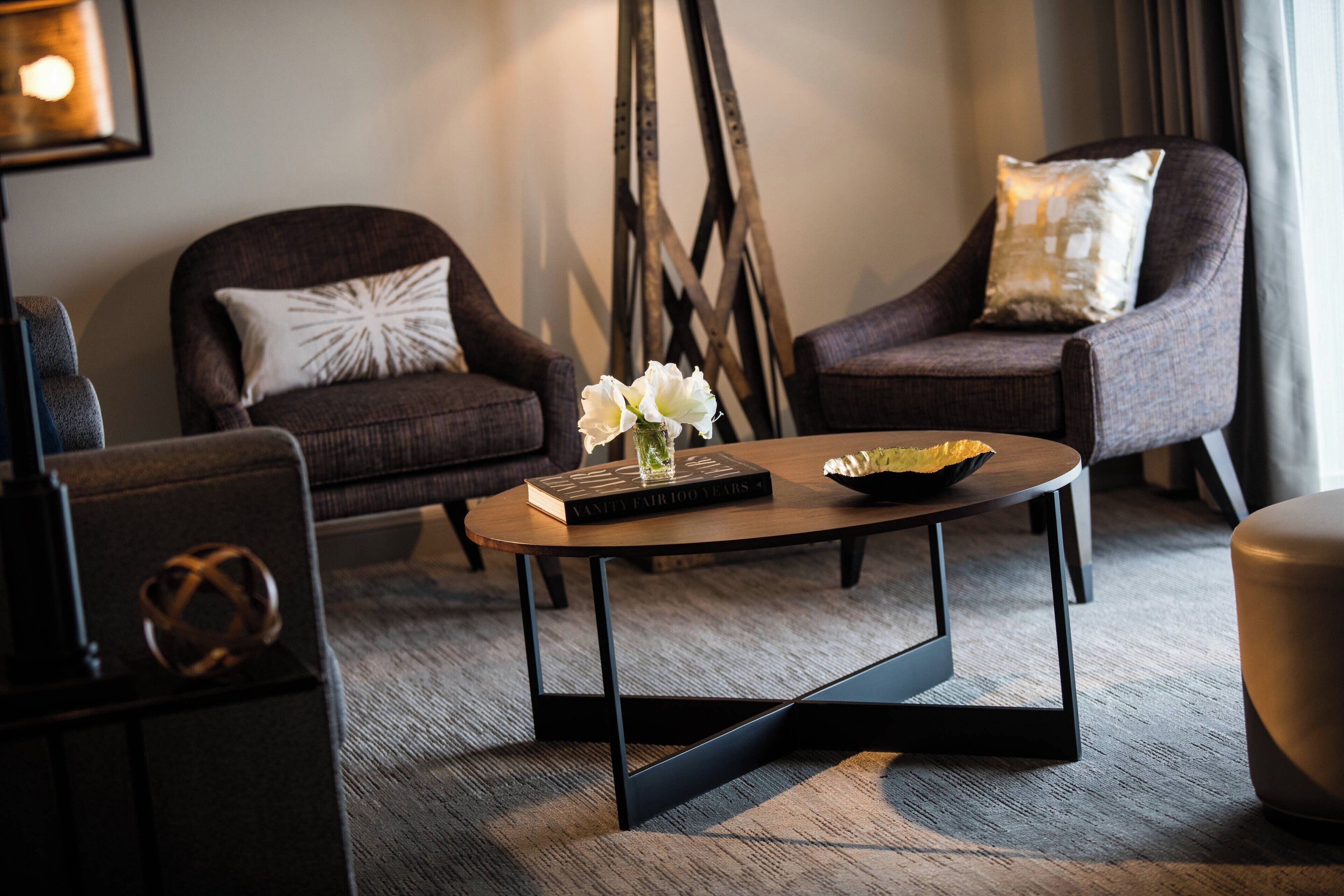 A modern living room with two dark armchairs, a round coffee table with a book and flowers, and a decorative wall piece.