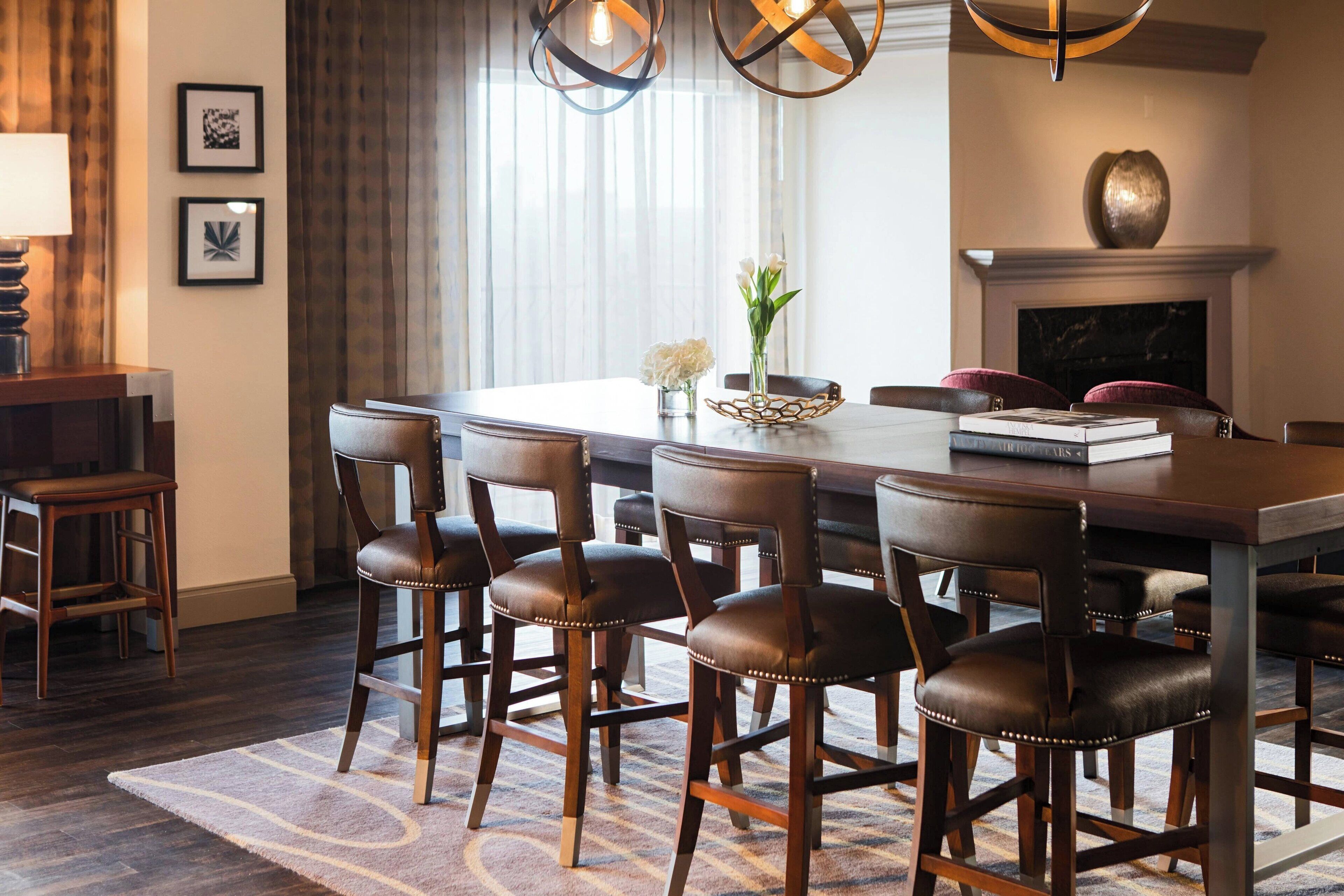 A dining area with a large wooden table, six leather chairs, a vase with flowers, and a fireplace.