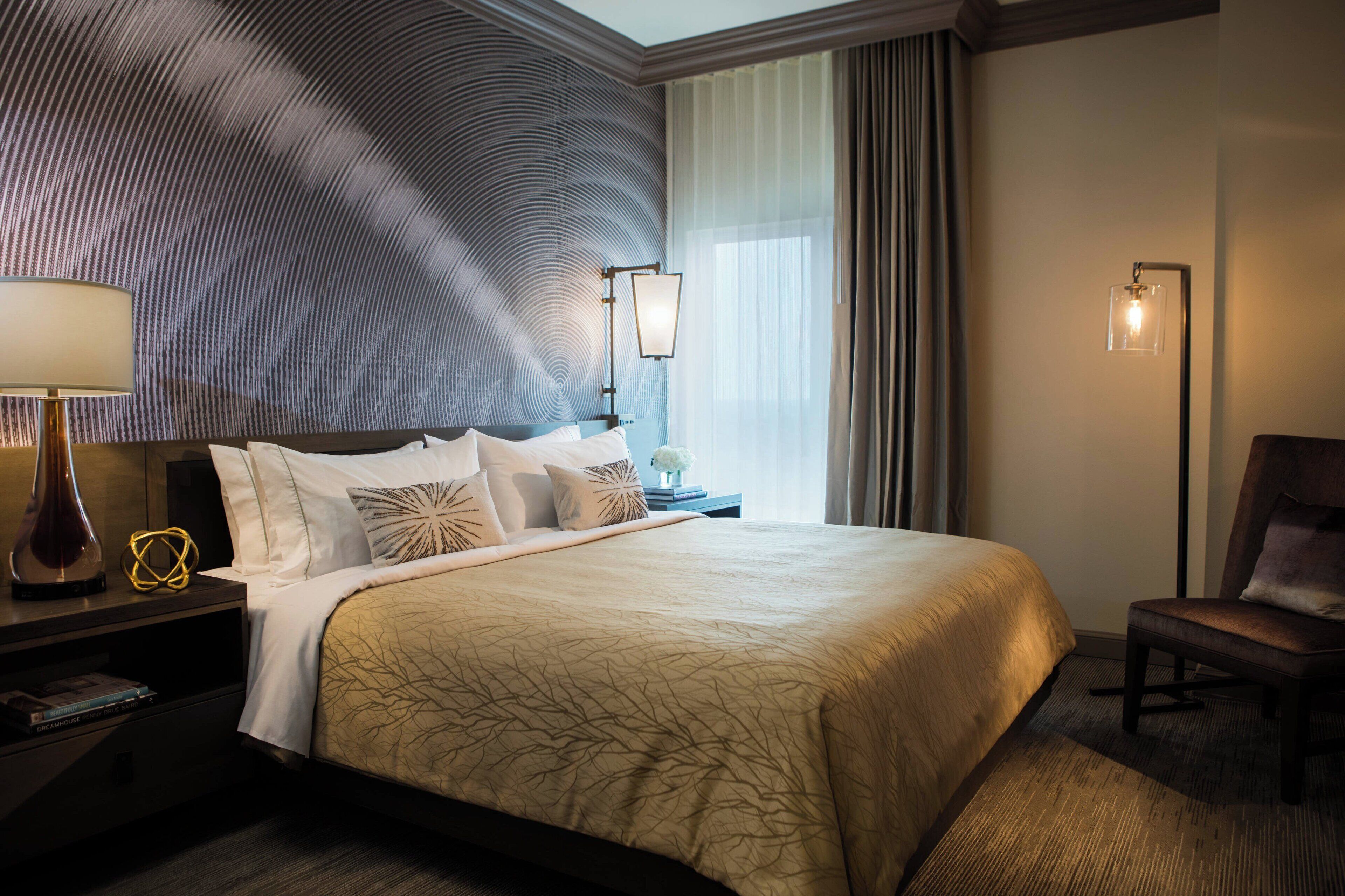 A hotel room with a large bed, a nightstand with a lamp, a chair, and a wall with a textured design.