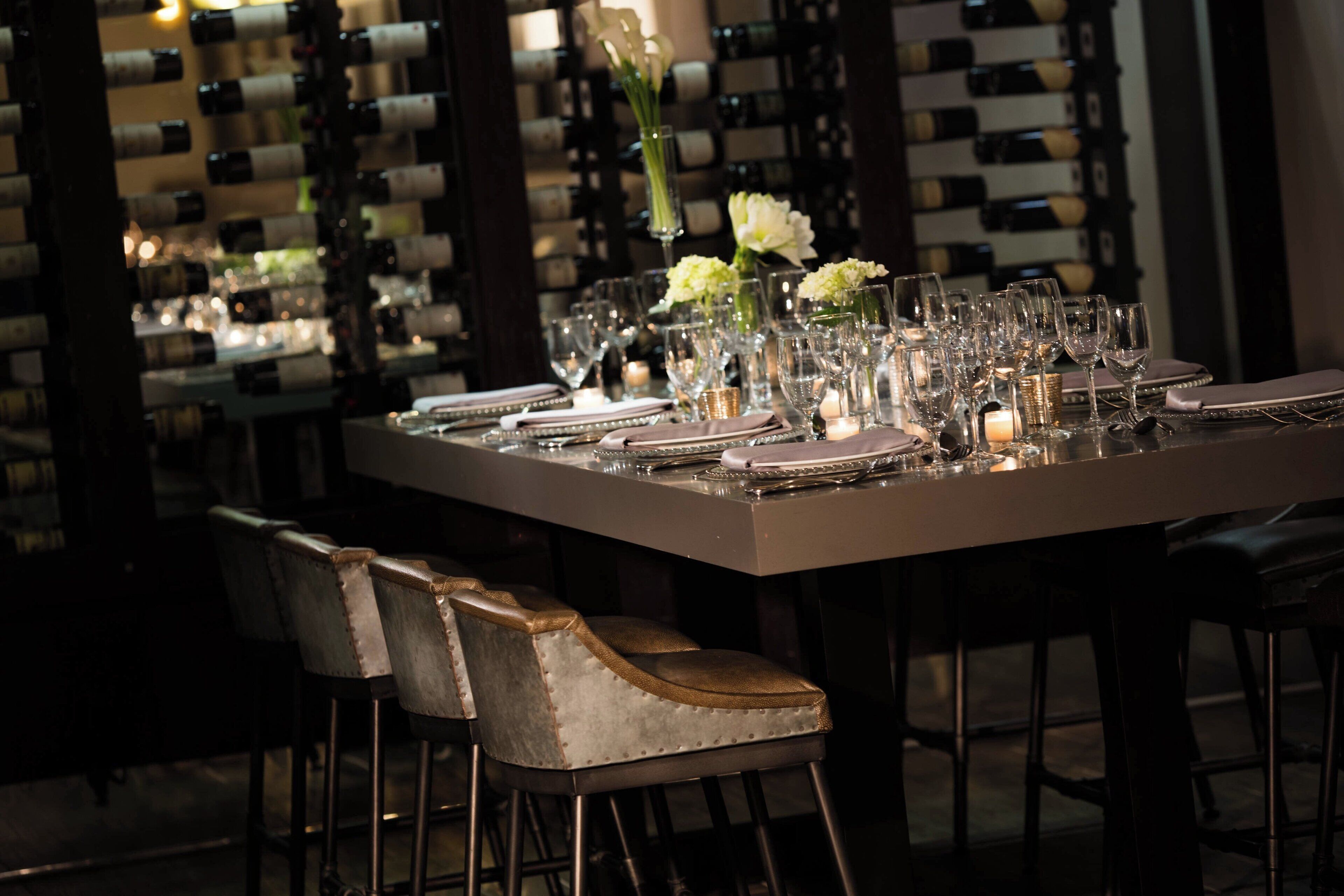 Mesa de restaurante posta com taças de vinho, pratos e talheres, com uma adega ao fundo.