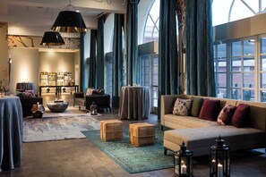 Amplio salón con un sofá grande, una mesita de centro y una estantería llena de libros y objetos decorativos. Hay dos ventanas grandes con cortinas, y una chimenea con repisa.