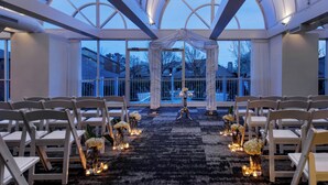 Hochzeitszeremonie mit weißen Stühlen und floralen Dekorationen unter einer großen Glasüberdachung mit natürlichem Licht.