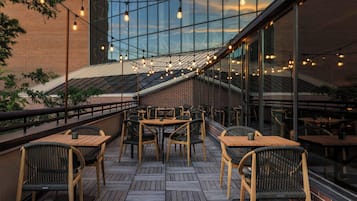 An outdoor seating area with wooden tables and chairs, a brick wall, and string lights.