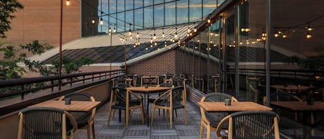 An outdoor seating area with wooden tables and chairs, a brick wall, and string lights.