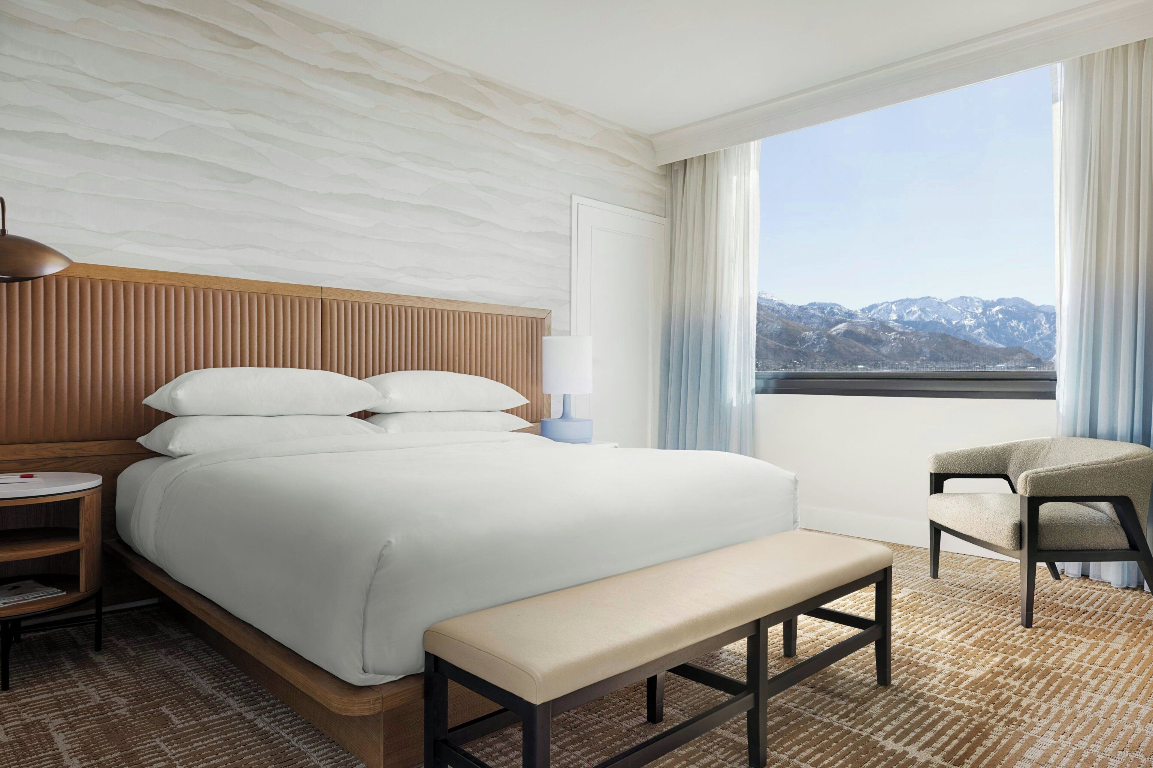 Une chambre d’hôtel avec un lit, une banquette, une chaise, une table de chevet et une vue sur les montagnes par la fenêtre.