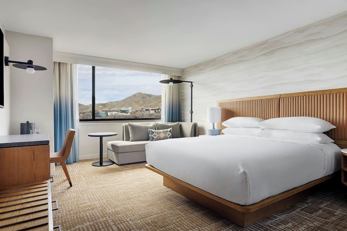 A hotel room with a bed, a desk, a chair, a small table, a lamp, and a view of the city.