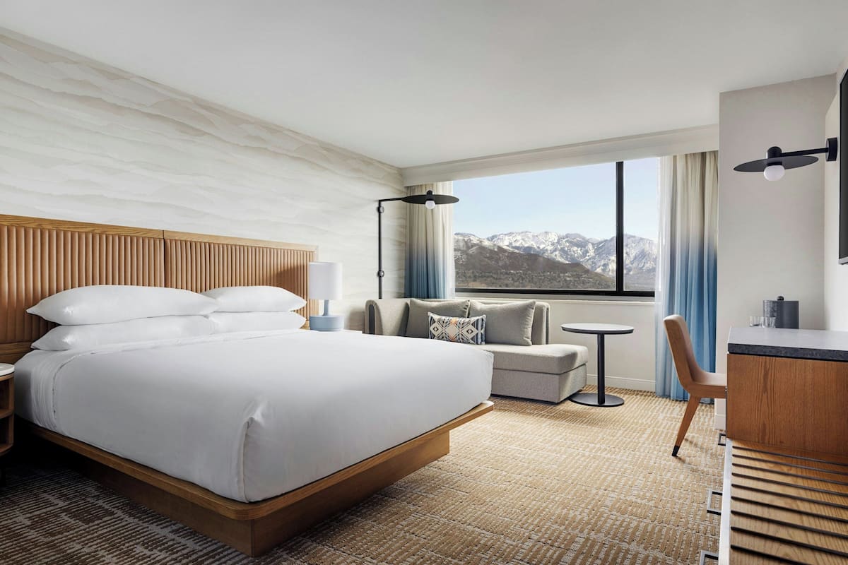 A hotel room with a bed, a sofa, a desk, a chair, a lamp, and a view of mountains.