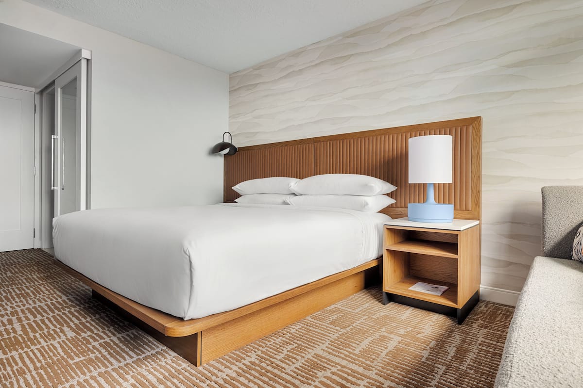 A hotel room with a wooden bed frame, white bedding, a nightstand with a lamp, and a patterned carpet.