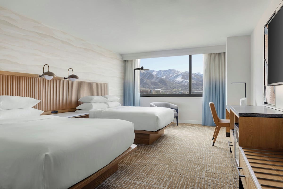 A hotel room with two beds, a desk, a chair, a large window with a mountain view, and a flat-screen TV.