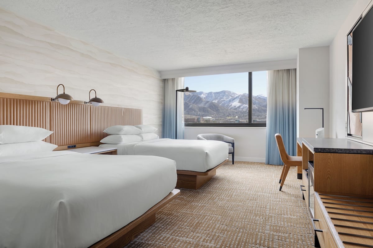 A hotel room with two beds, a desk, a chair, a TV, and a large window with a mountain view.