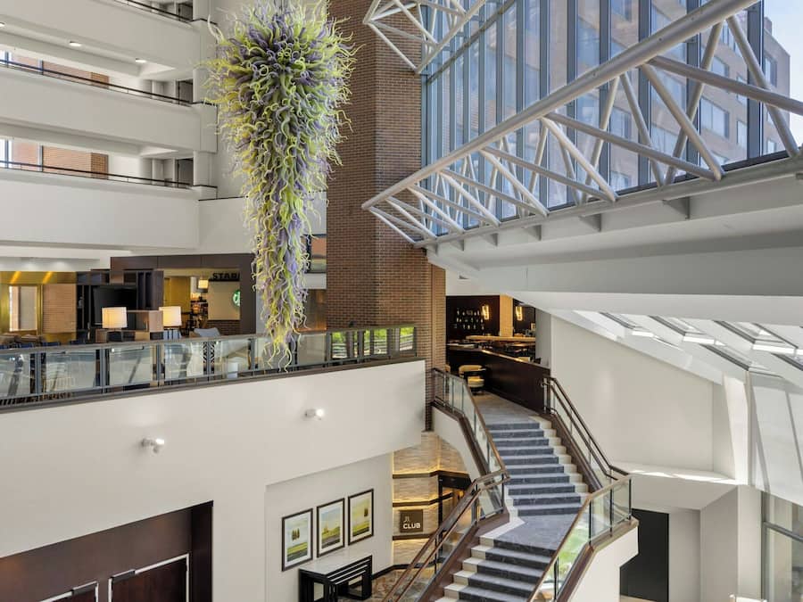Un vestíbulo de hotel de varios niveles con una escalera central, una instalación de plantas colgantes y una estructura moderna de techo de cristal.