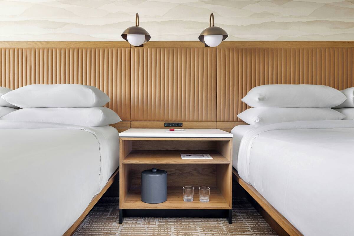 Hotelkamer met twee bedden, een houten hoofdbord, een nachtkastje met een lamp, een klein plankje met glazen en een prullenbak.