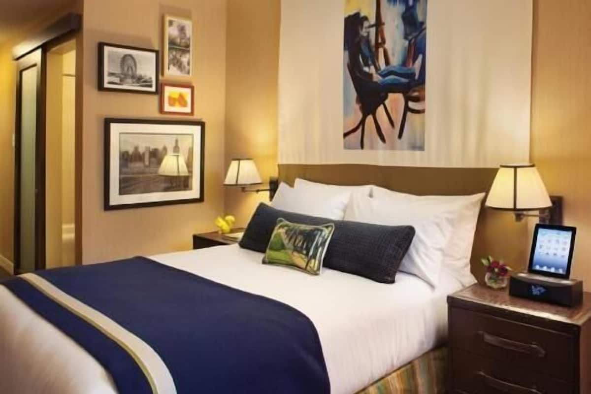 Habitación de hotel con una cama, mesitas de noche, lámpara, reloj digital y cuadros en la pared.