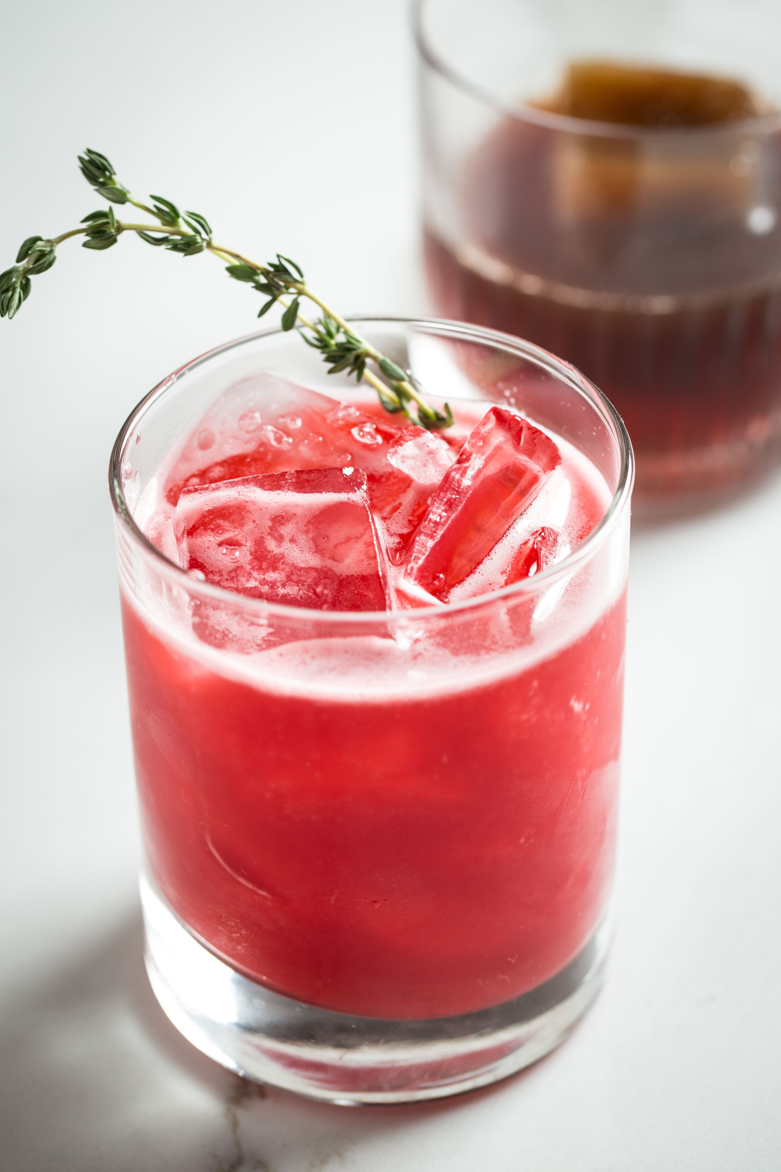 A glass of red beverage with ice cubes and a sprig of thyme.